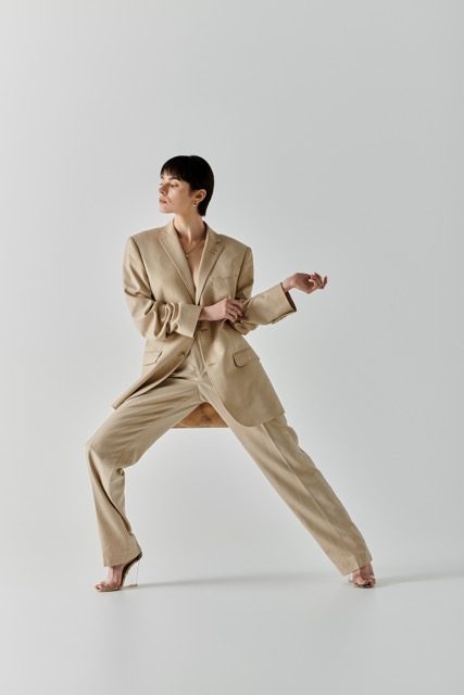 A woman in a beige suit poses with her leg extended in a studio.