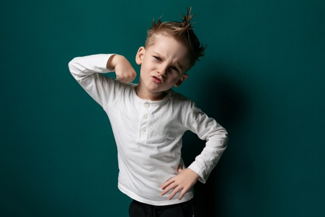 A young boy is standing still, smiling, and posing in front of the camera for a photograph. He appears confident and comfortable in his posture as the photographer captures his image.