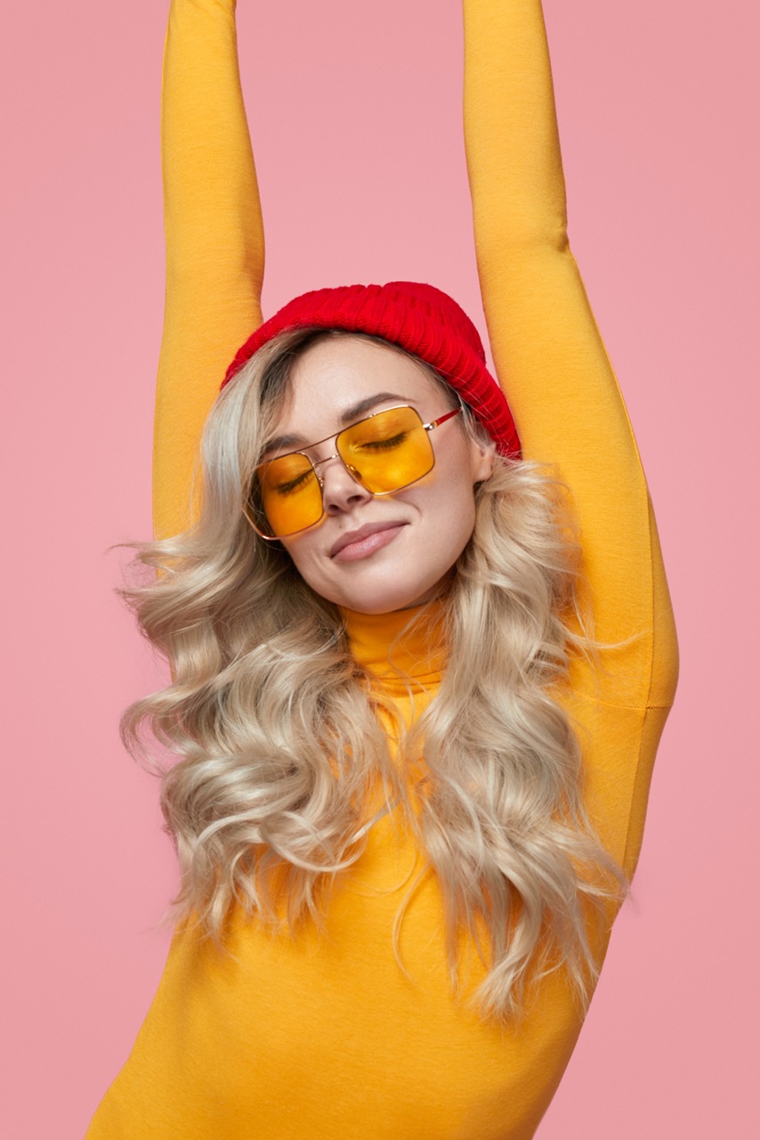 Blond young female in yellow turtleneck and trendy red hat closing eyes and stretching raised arms against pink background in morning